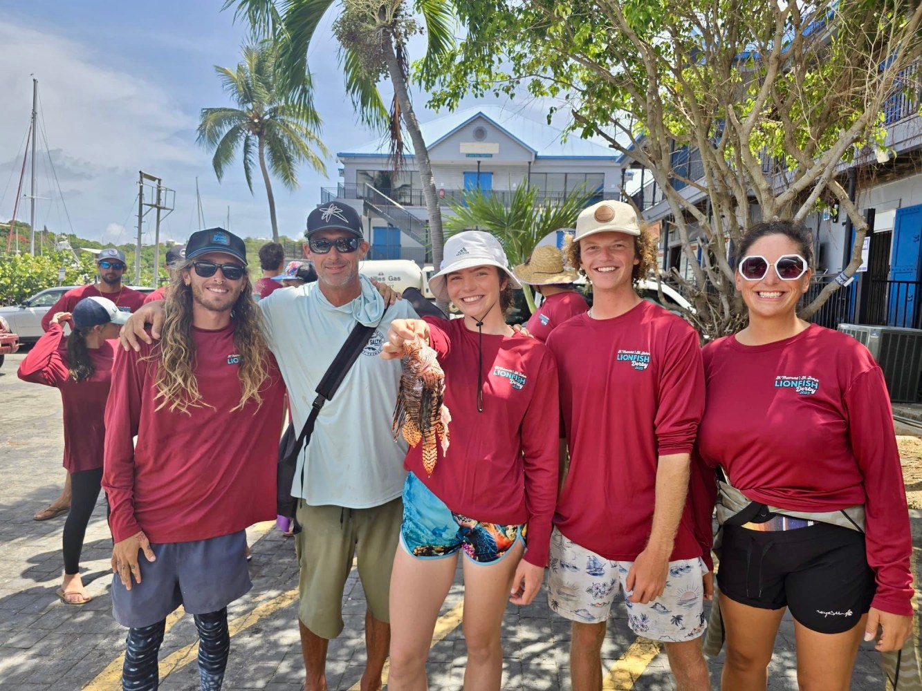 Group of people in matching red shirts posing outdoors with a fish, tropical setting with palm trees.