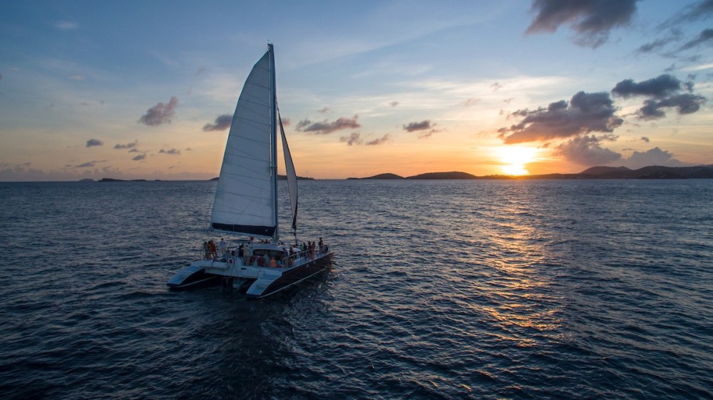 Sailboat on ocean at sunset with cloudy sky and distant islands visible.