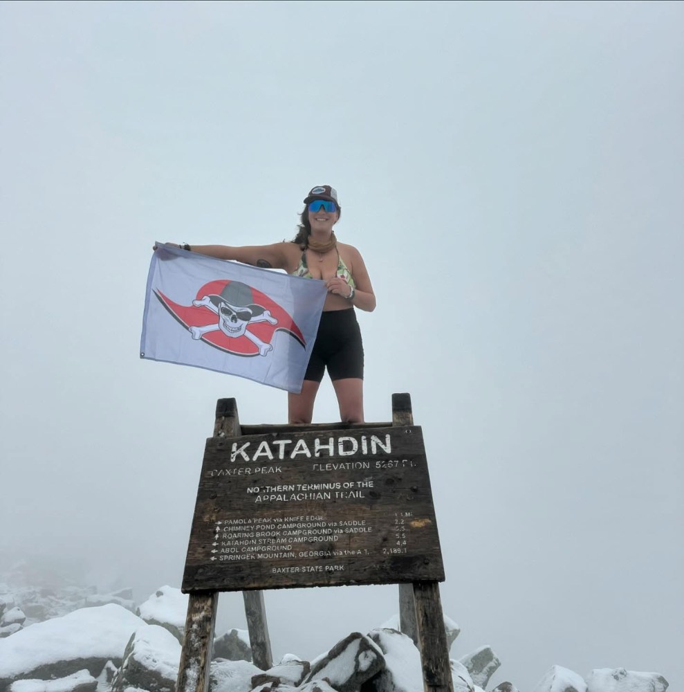 Person standing on Katahdin sign, holding a flag with a skull emblem on a foggy day.