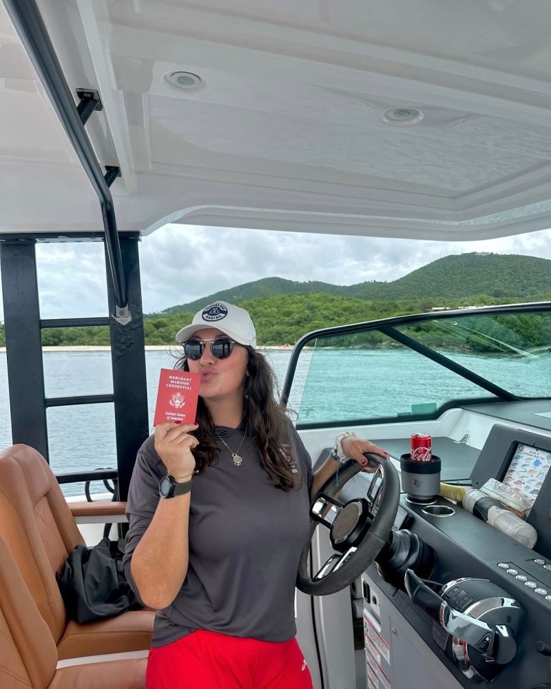 Person in a cap holds a red book while steering a boat with scenic hills in the background.