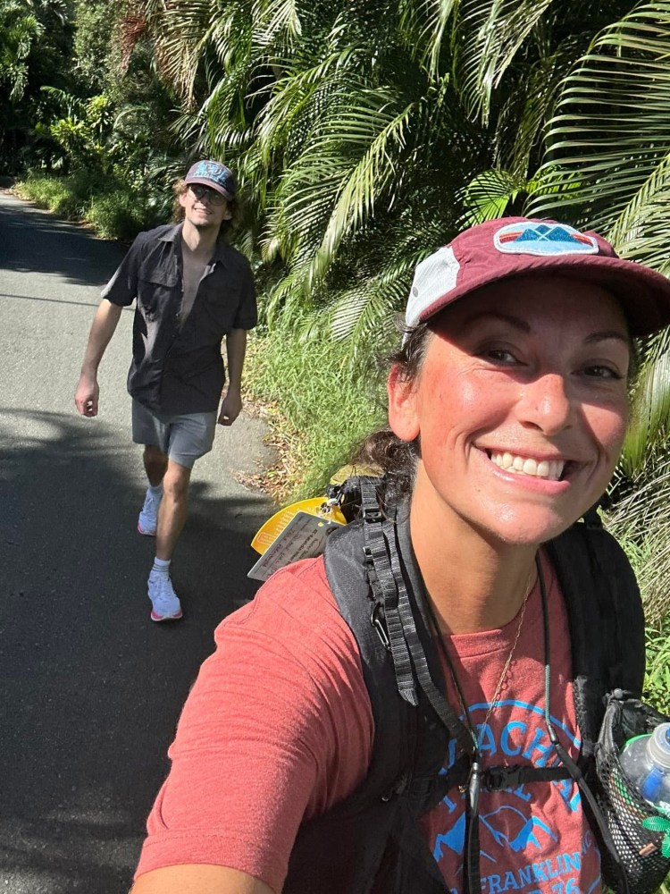 Two people walking on a path with tropical plants, one smiling for a selfie.