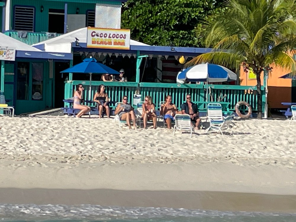 People relaxing on beach chairs by a bar with palm trees and beach umbrellas.