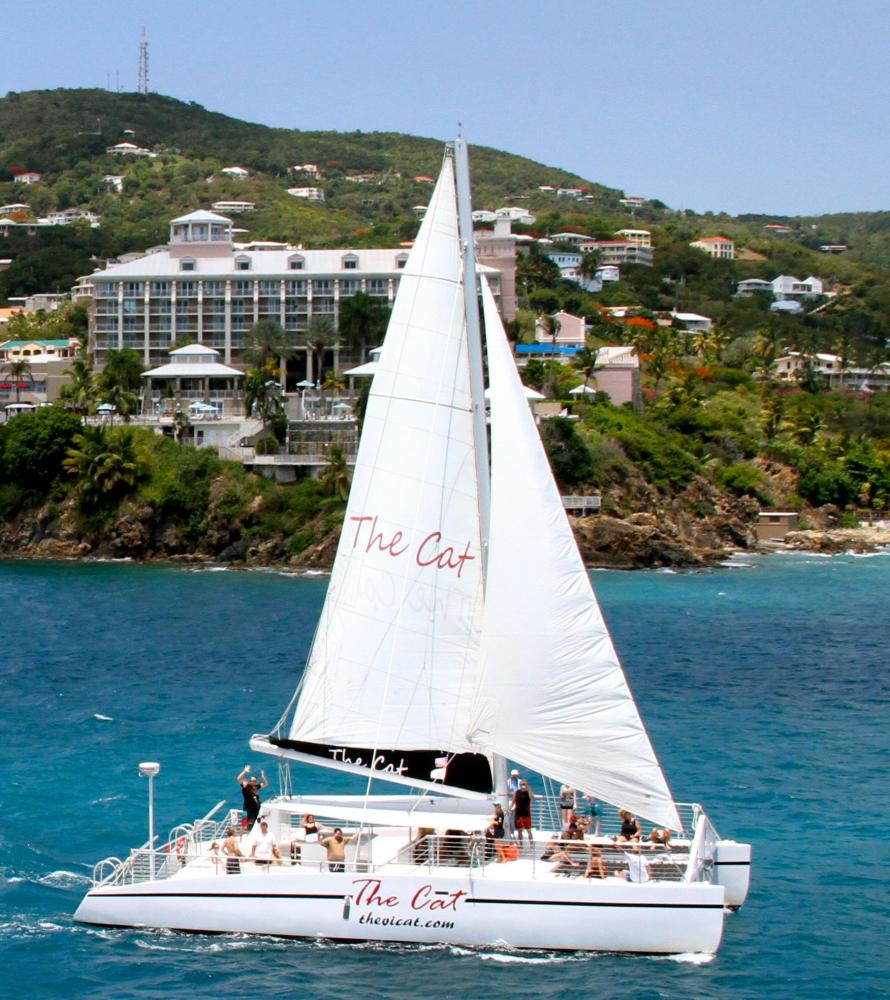 Catamaran with people sailing near a tropical shoreline with buildings and greenery.
