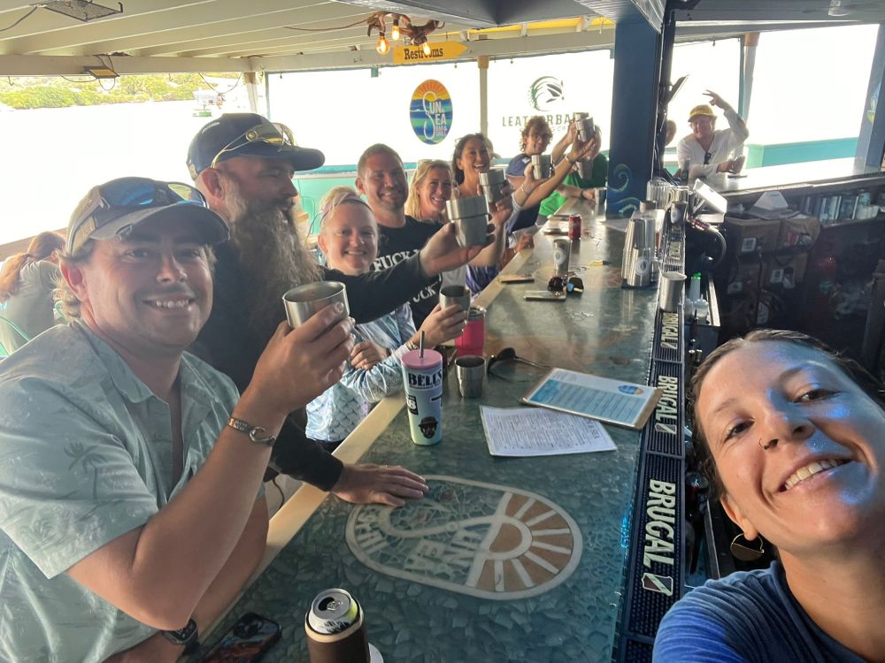 Group of people smiling and holding drinks at a bar, woman taking selfie in the foreground.