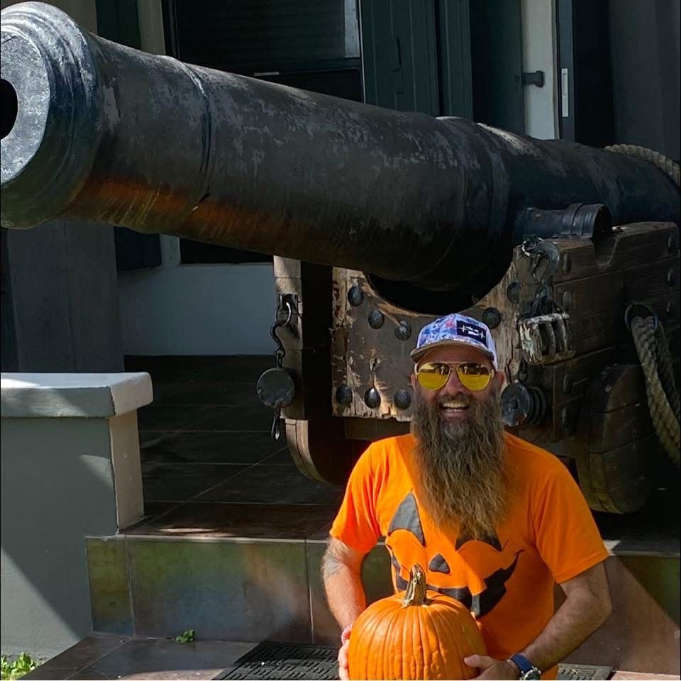 3f97191c-2d3b-4554-9cf1-f88ff3b22625 Man in pumpkin shirt holds pumpkin in front of large cannon.
