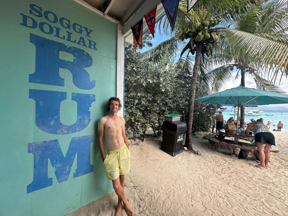 Person in yellow shorts leans on 'Soggy Dollar Rum' wall at beach with palm trees and people nearby.