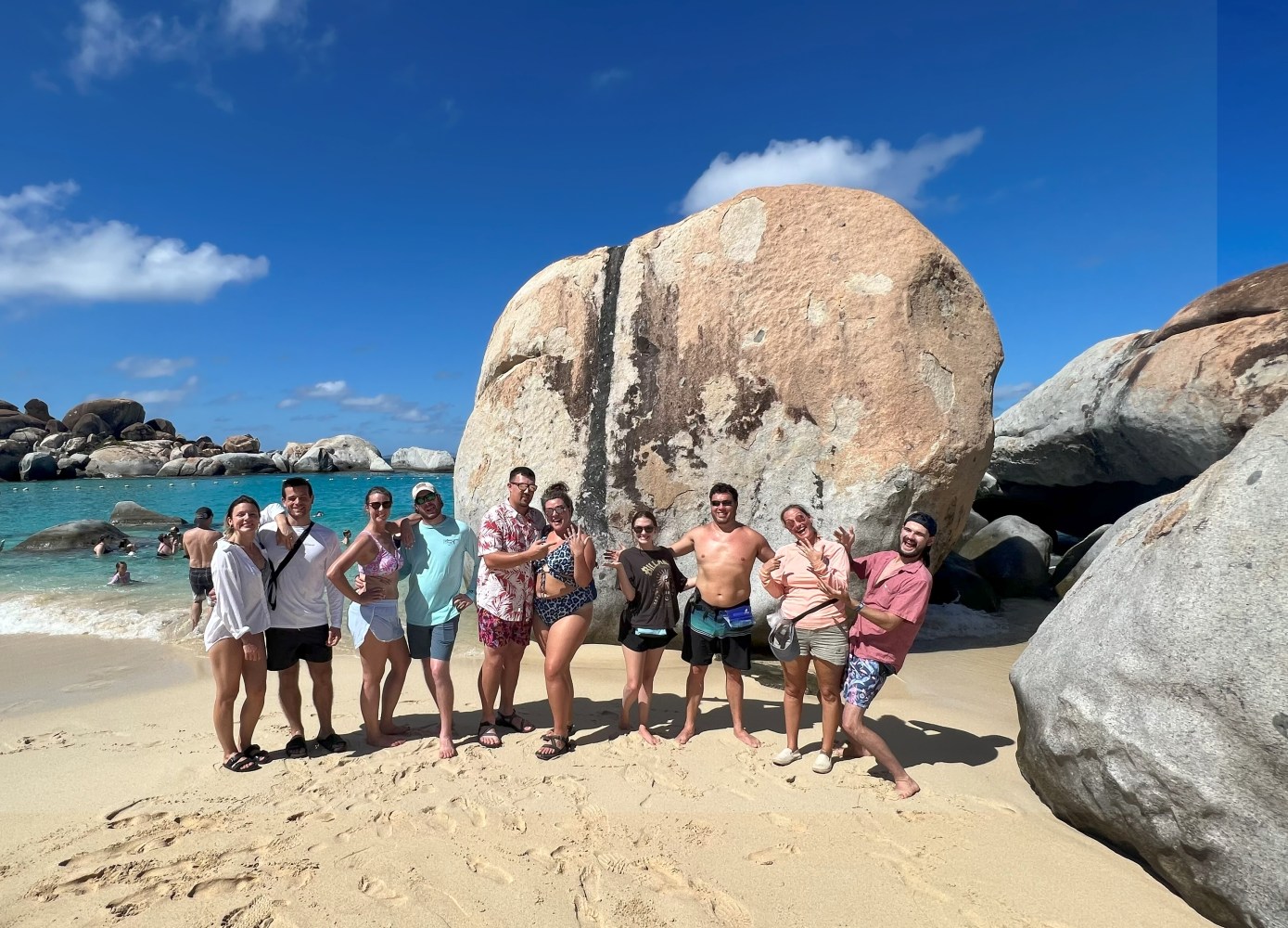 Group of people posing on a sandy beach with large rocks and clear blue sky.