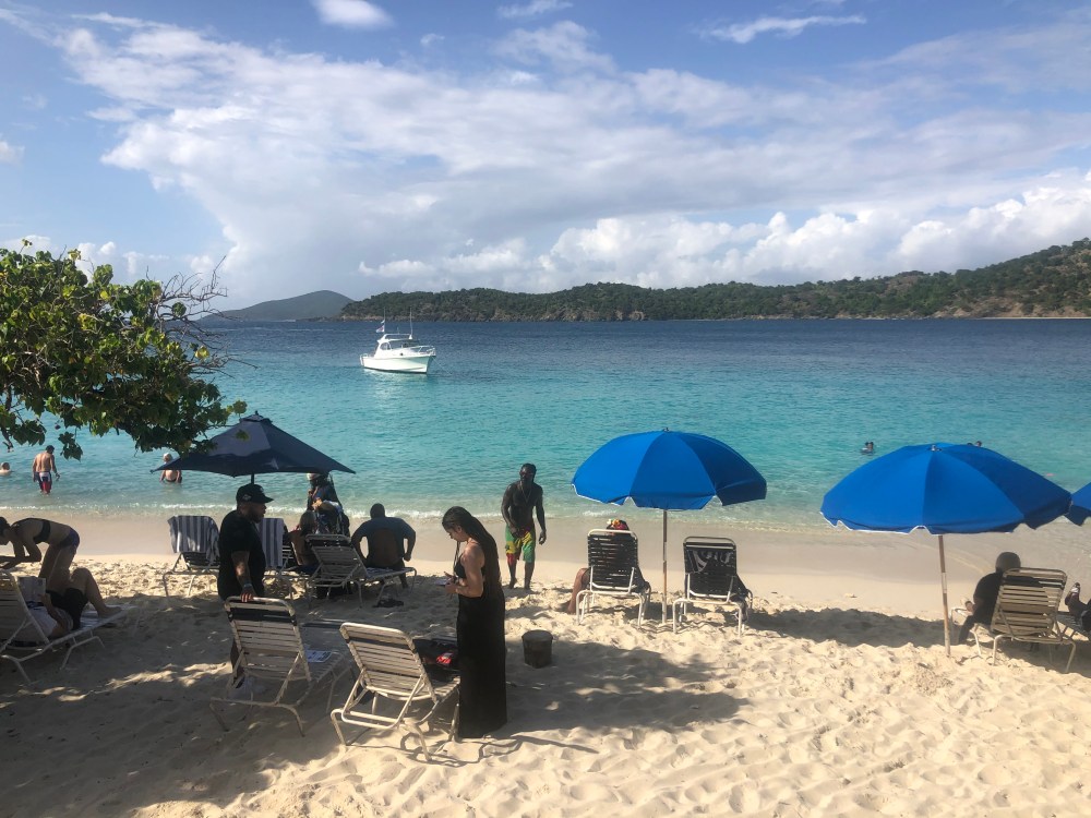 Beach scene with people, blue umbrellas, and a boat on clear water under a partly cloudy sky.