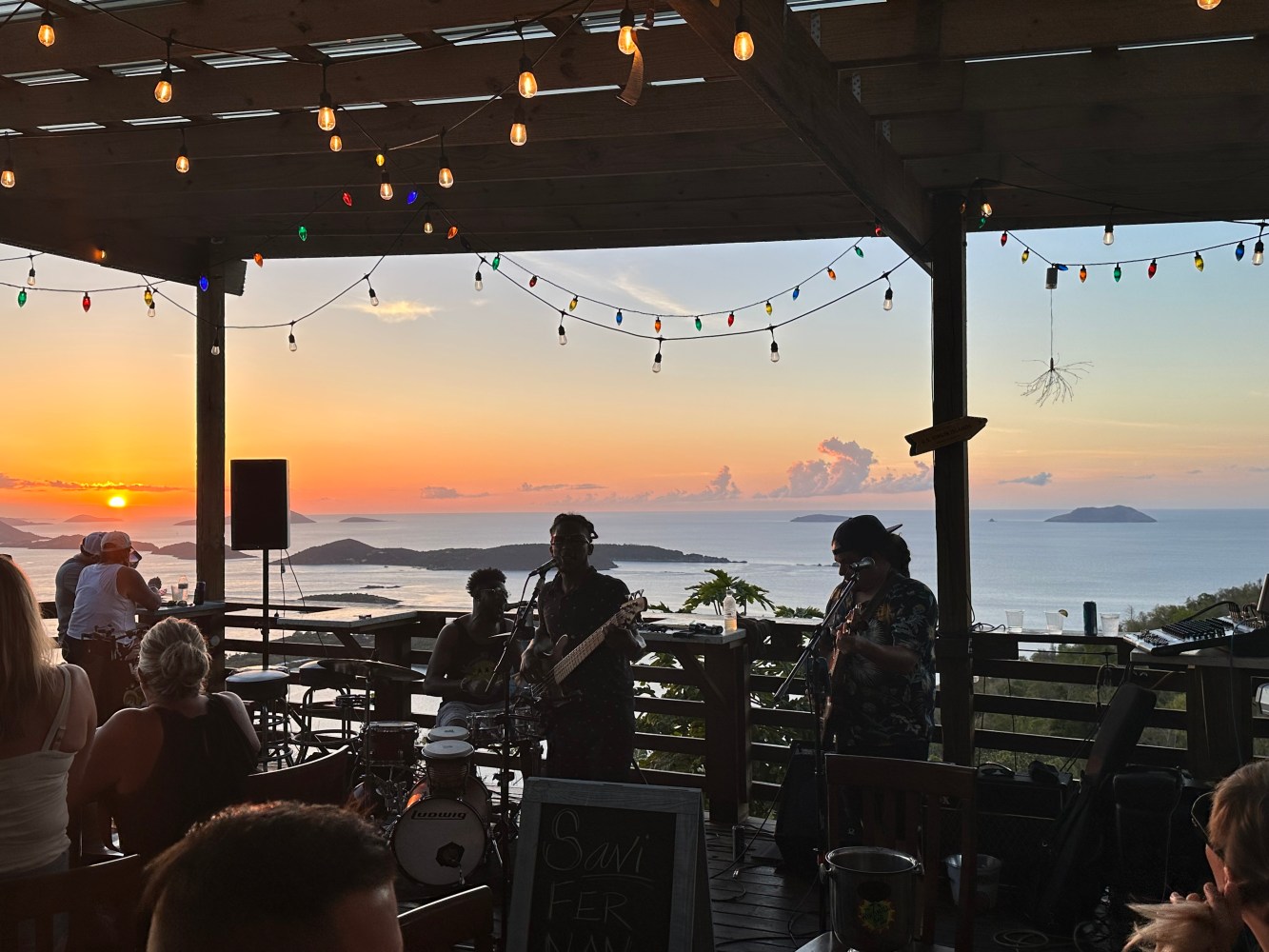 Musicians perform at a beachside venue during a vibrant sunset, with string lights overhead.
