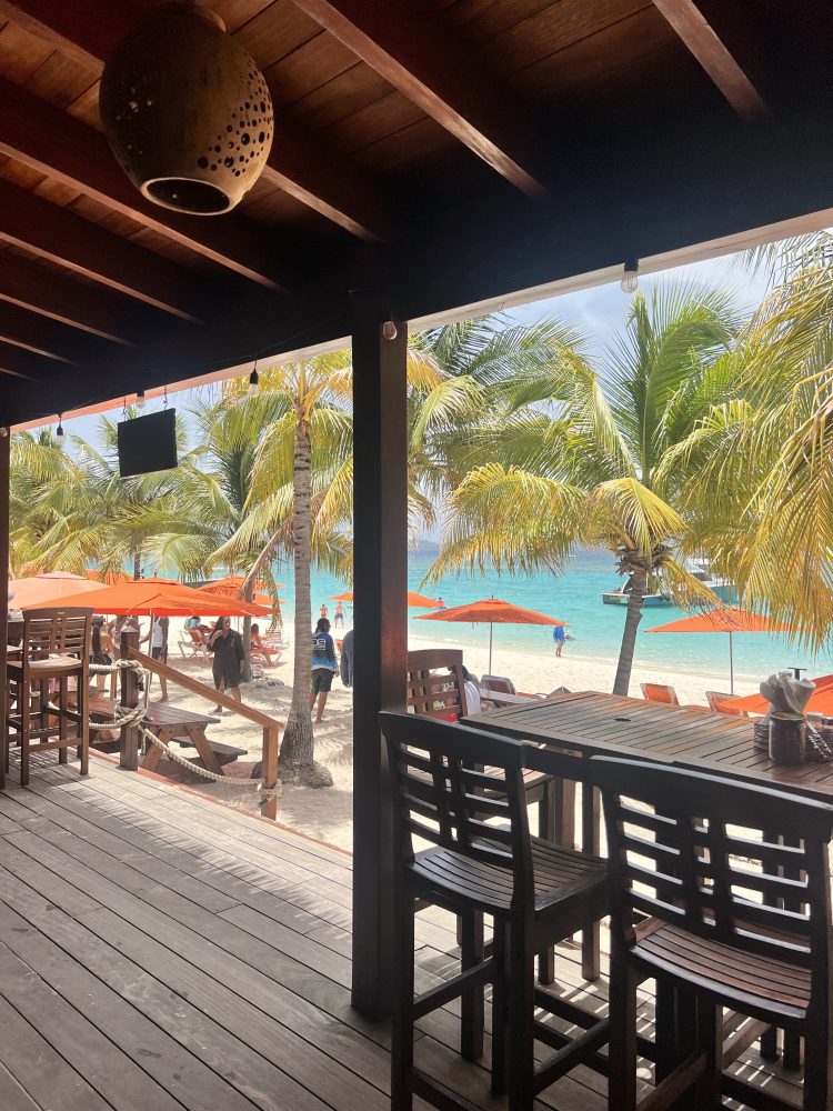 Beachside view from a wooden terrace with bar chairs, palm trees, orange umbrellas, and turquoise sea.