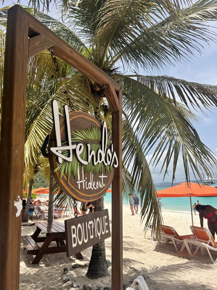 Beach scene with 'Hendo's Hideout' sign under palm trees, orange umbrellas and lounge chairs.