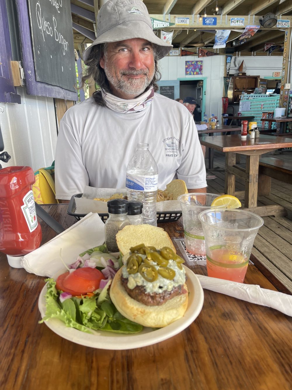 Smiling man with hat at table with jalapeño burger, salad, and drinks in open-air restaurant.