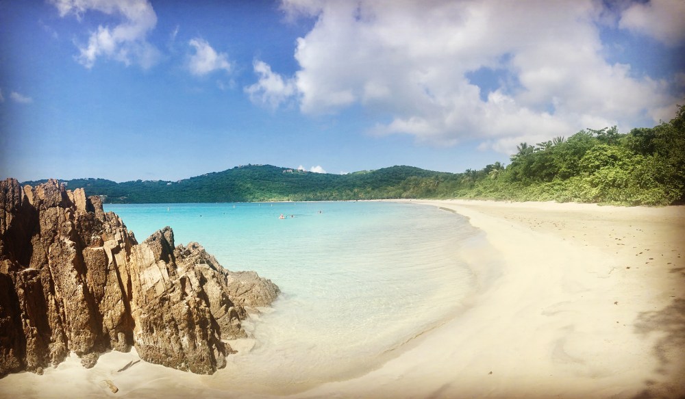 Scenic beach with turquoise water, rocky foreground, sandy shore, and lush green hills under a blue sky.
