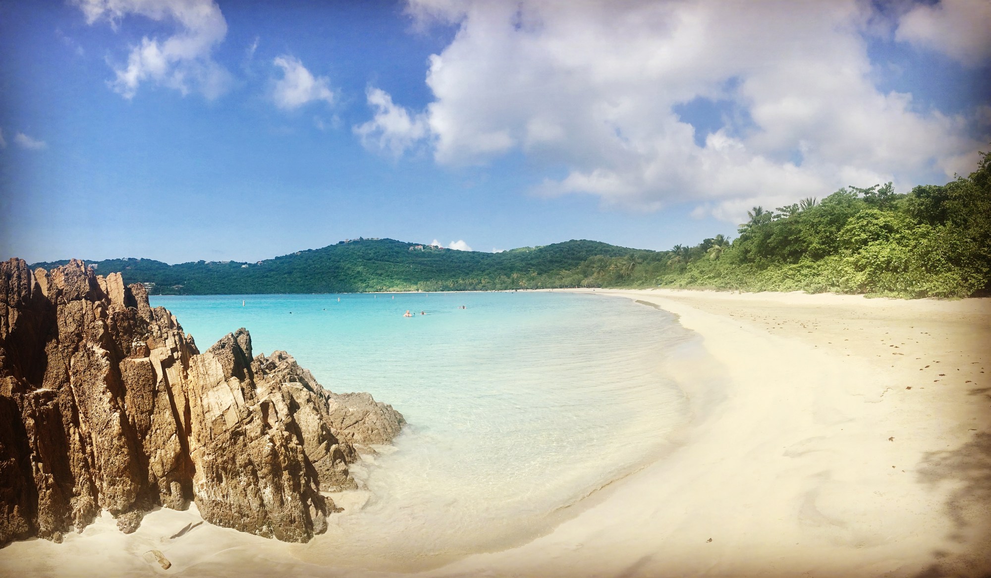 Magens Bay Scenic beach with turquoise water, rocky foreground, sandy shore, and lush green hills under a blue sky.