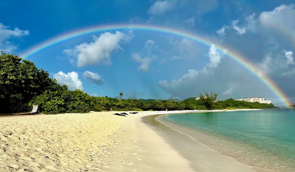 Sandy beach with lush greenery and a bright rainbow arching across a blue sky above calm turquoise waters.