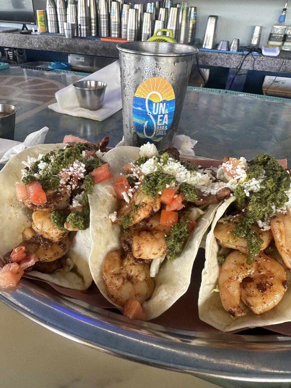 Three shrimp tacos with toppings next to a branded metal cup on a bar counter.