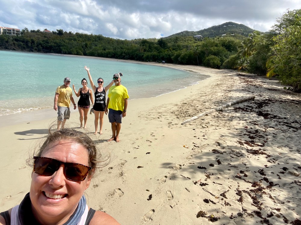 Group of people walking on a sandy beach with turquoise water and trees in the background.