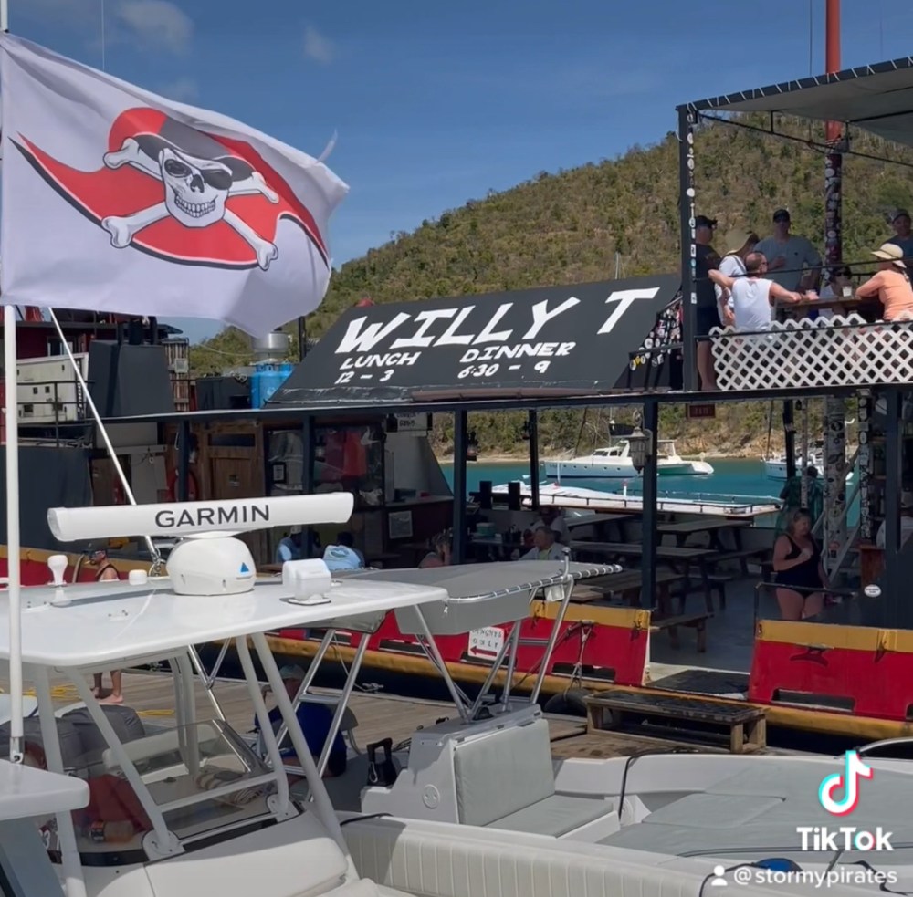 Floating bar with pirate flag, named Willy T, near a boat, with people dining and hilly island background.