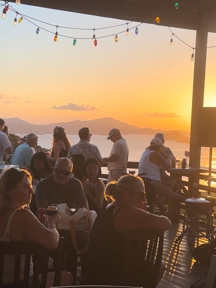 People at an outdoor bar during sunset with colorful string lights overhead.