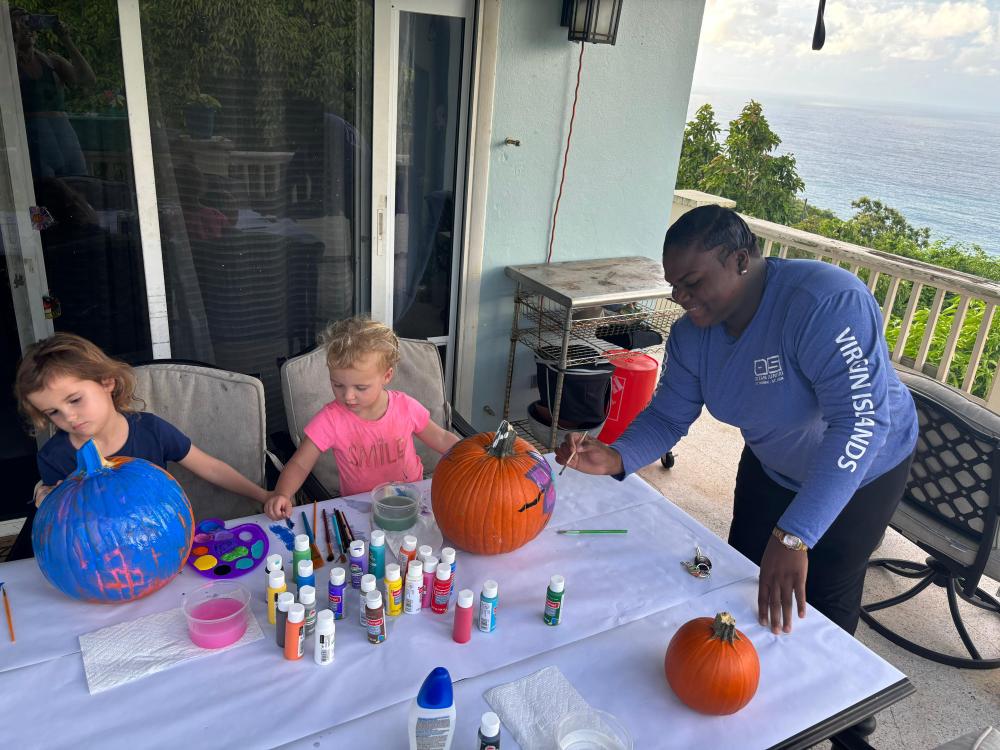 Three people painting pumpkins on a table, outdoors with an ocean view.