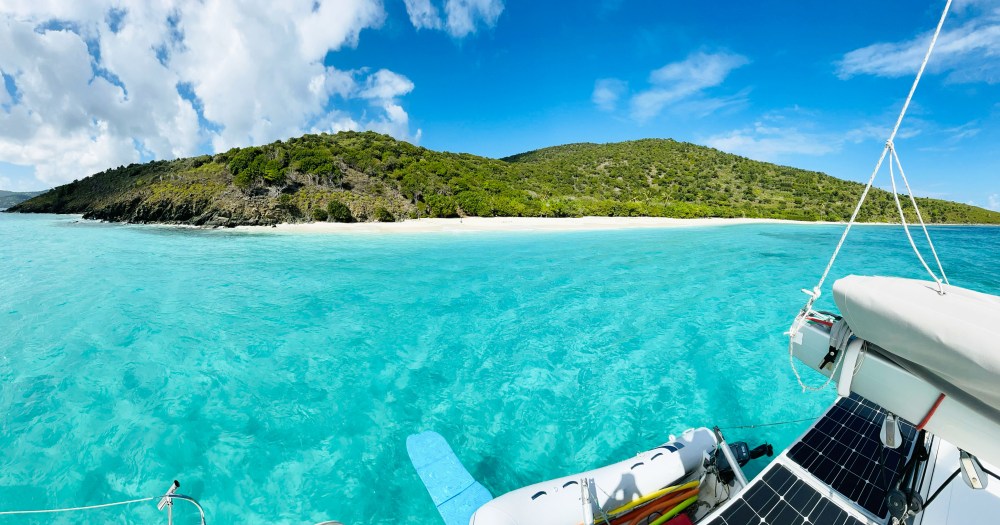 Sailboat on clear turquoise water near a green, hilly island with a sandy beach under a blue sky with clouds.