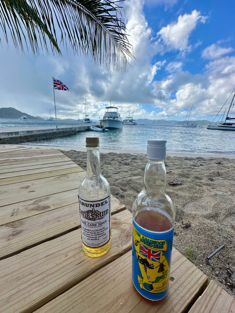 Two rum bottles on a wooden deck by the ocean with boats and a Union Jack flag in the background.