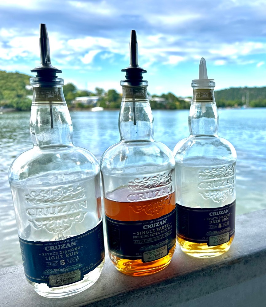 Three rum bottles with pourers on a ledge by a lake with trees and clouds in the background.