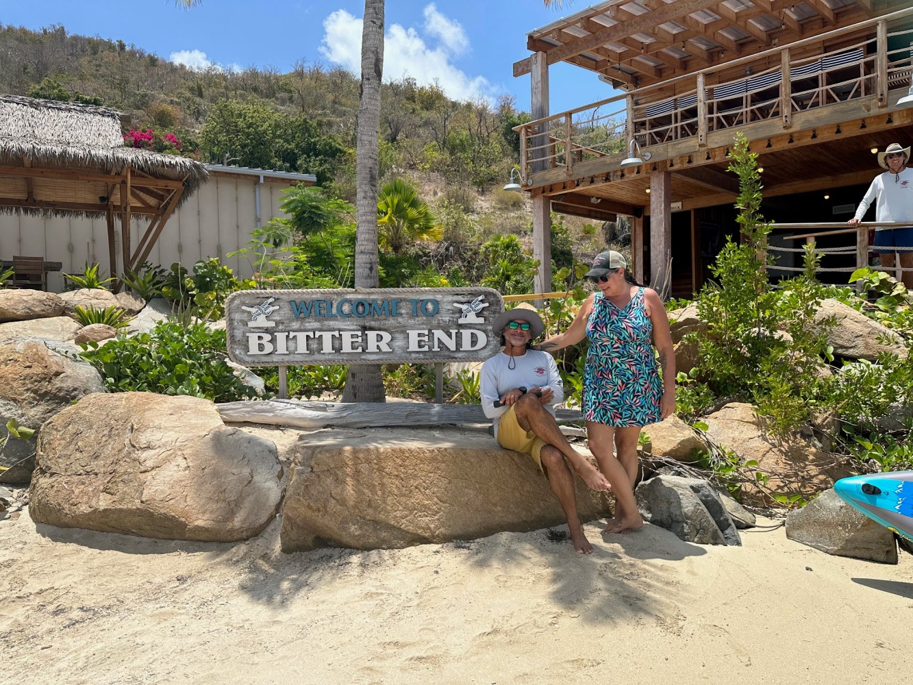 Two people sitting by 'Welcome to Bitter End' sign on a sunny beach.