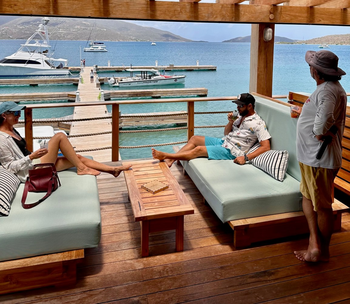 Three people relax on a wooden deck by the sea, with boats in the background.