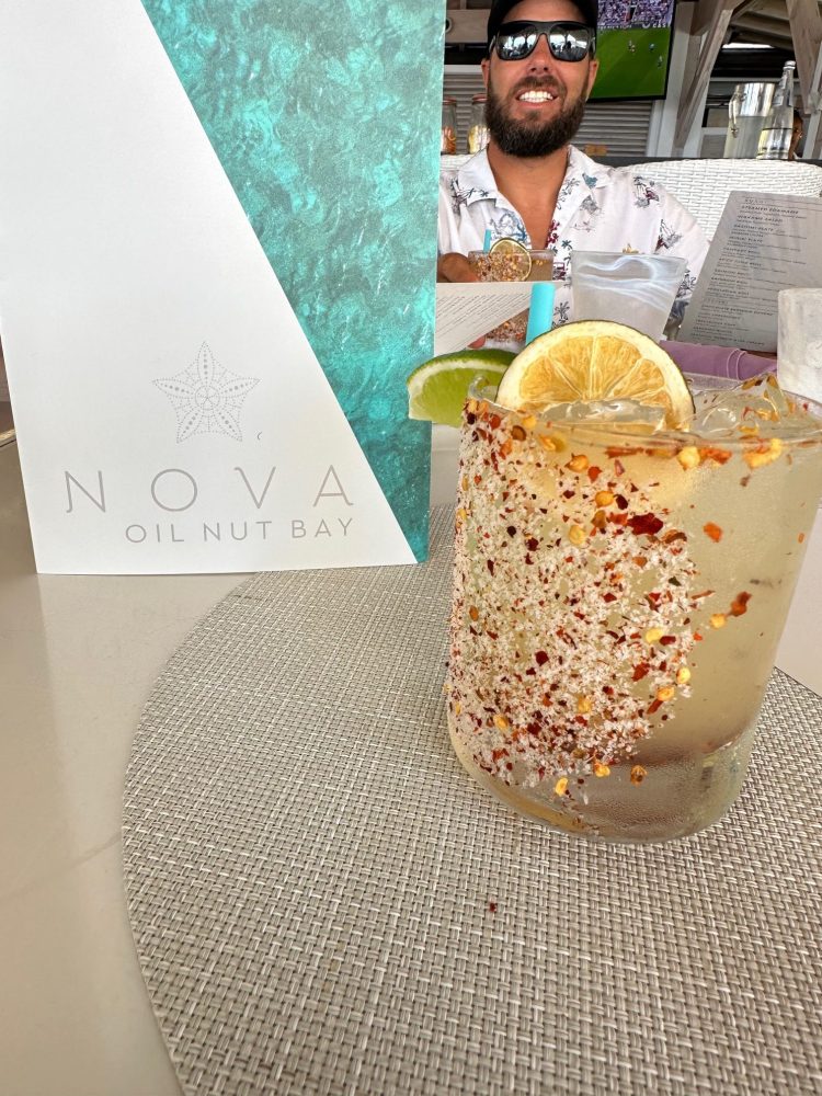 Spicy cocktail with lemon and lime, menu and smiling man in background at NOVA Oil Nut Bay.