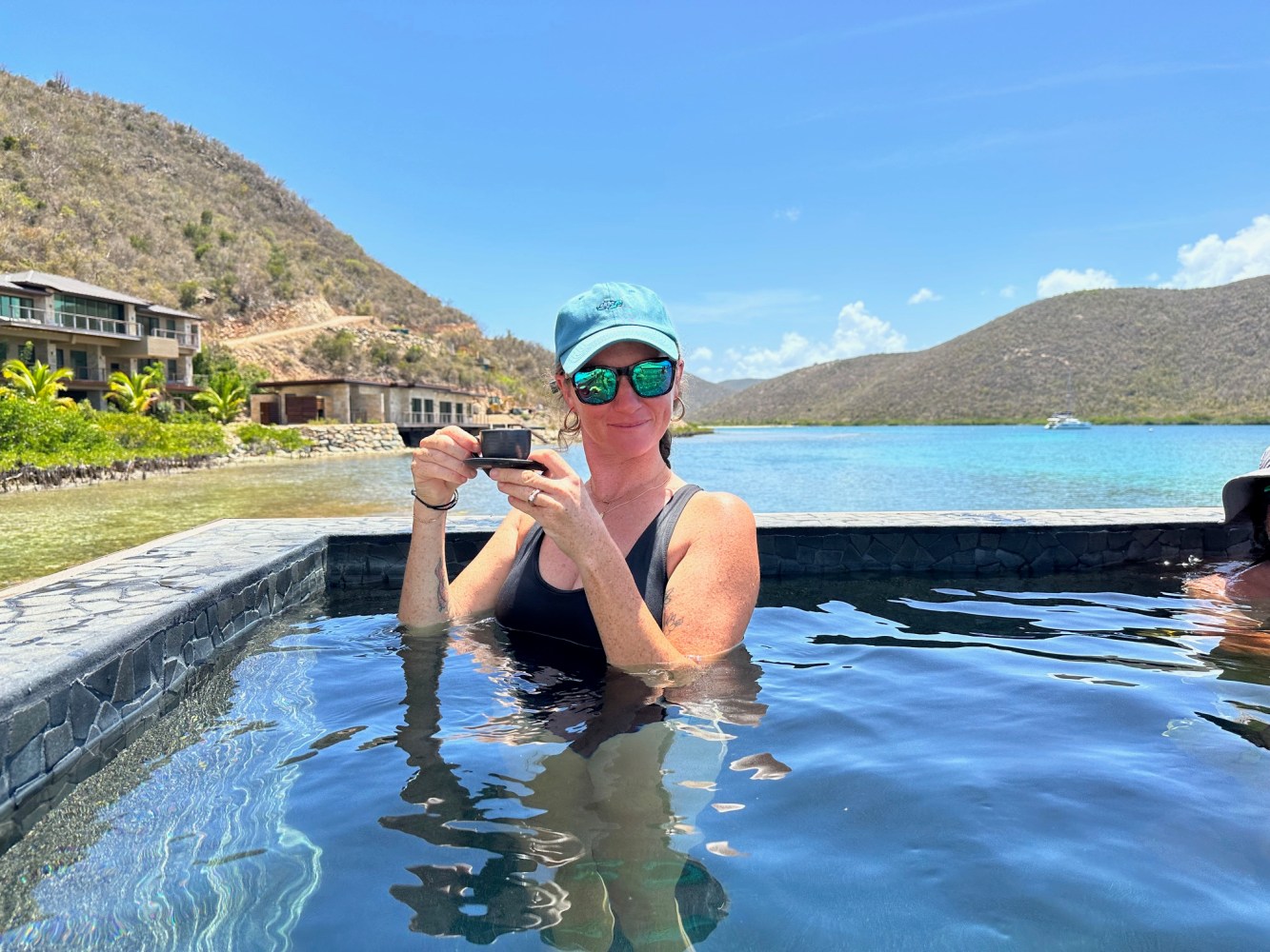Person in a cap holding a camera in a pool with a scenic view of hills and water.