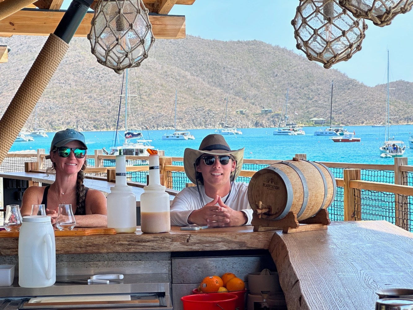 Two people at a seaside bar with boats on blue water and hills in the background.