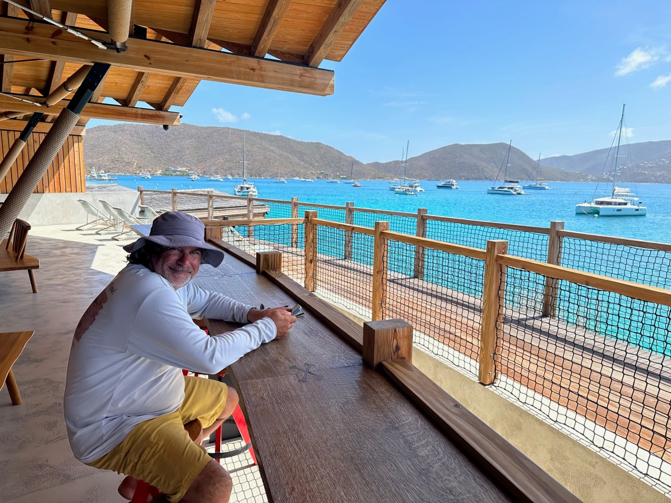 Person sitting at wooden counter overlooking a bay with boats and mountains.