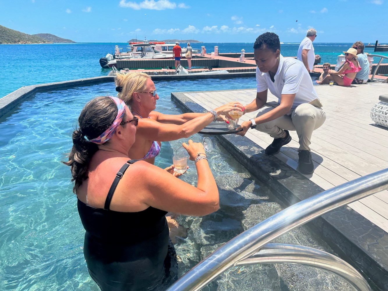 Two women in a pool receive drinks from a server on a deck by the ocean.