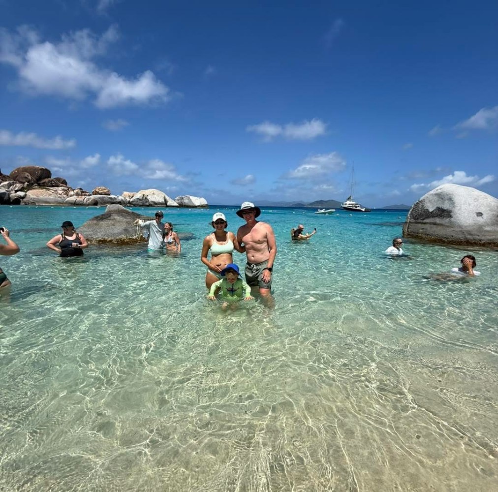 People in shallow clear water with boulders and a boat in the background under a sunny sky.