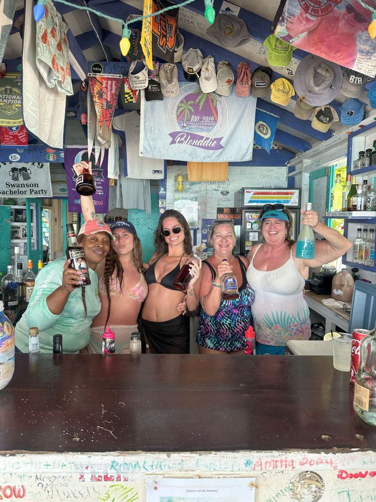 Five people smiling and holding bottles in a decorated bar with hats and shirts hanging above.