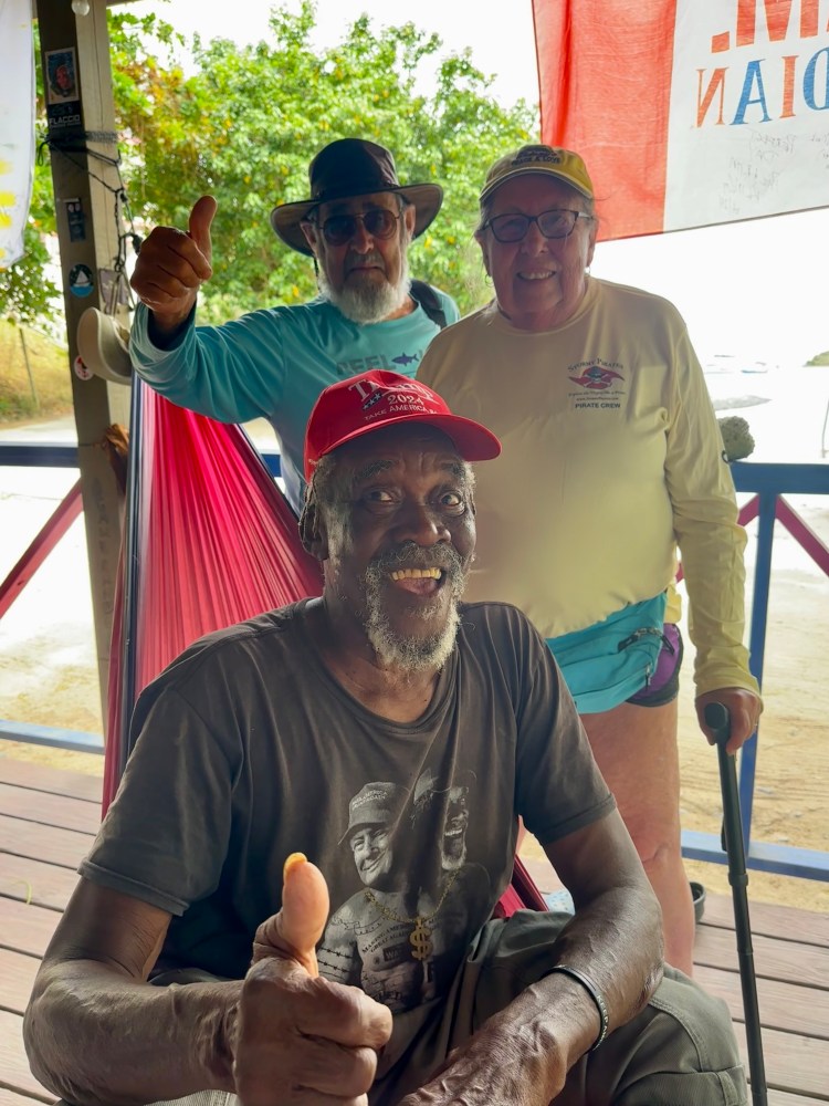 Three smiling people give thumbs up, one seated, on a wooden terrace with trees in the background.