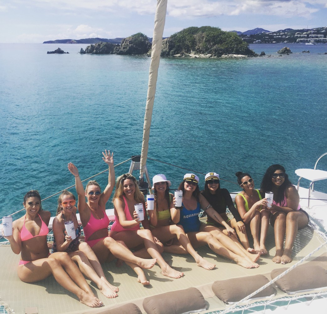 Group of women in swimsuits on a boat, holding drinks, with ocean and islands in the background.