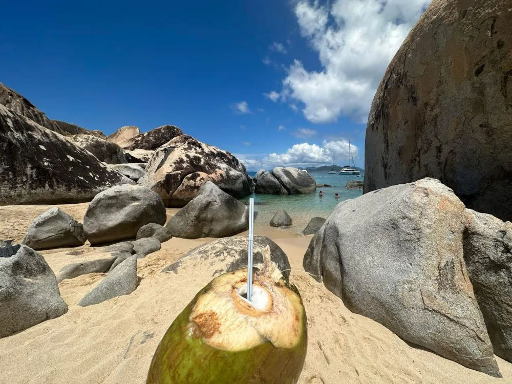 View of a tropical beach with large rocks and a coconut with a straw in the foreground.