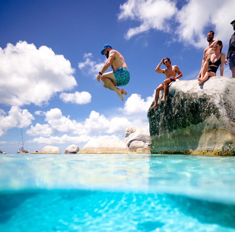 Man in blue shorts jumping off rocks into clear water, with friends watching and cloudy sky above.