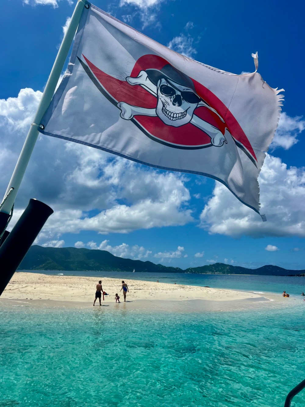 Pirate flag on boat near a sandy beach with people and clear blue water.