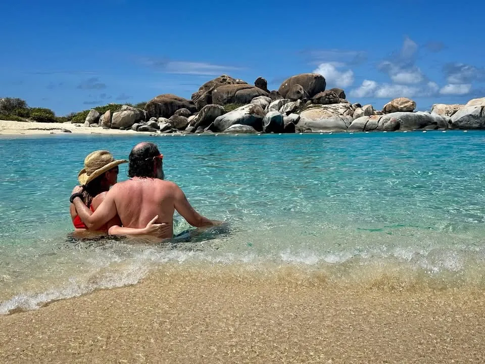 Couple in water facing rocks, bright day, clear blue sea.