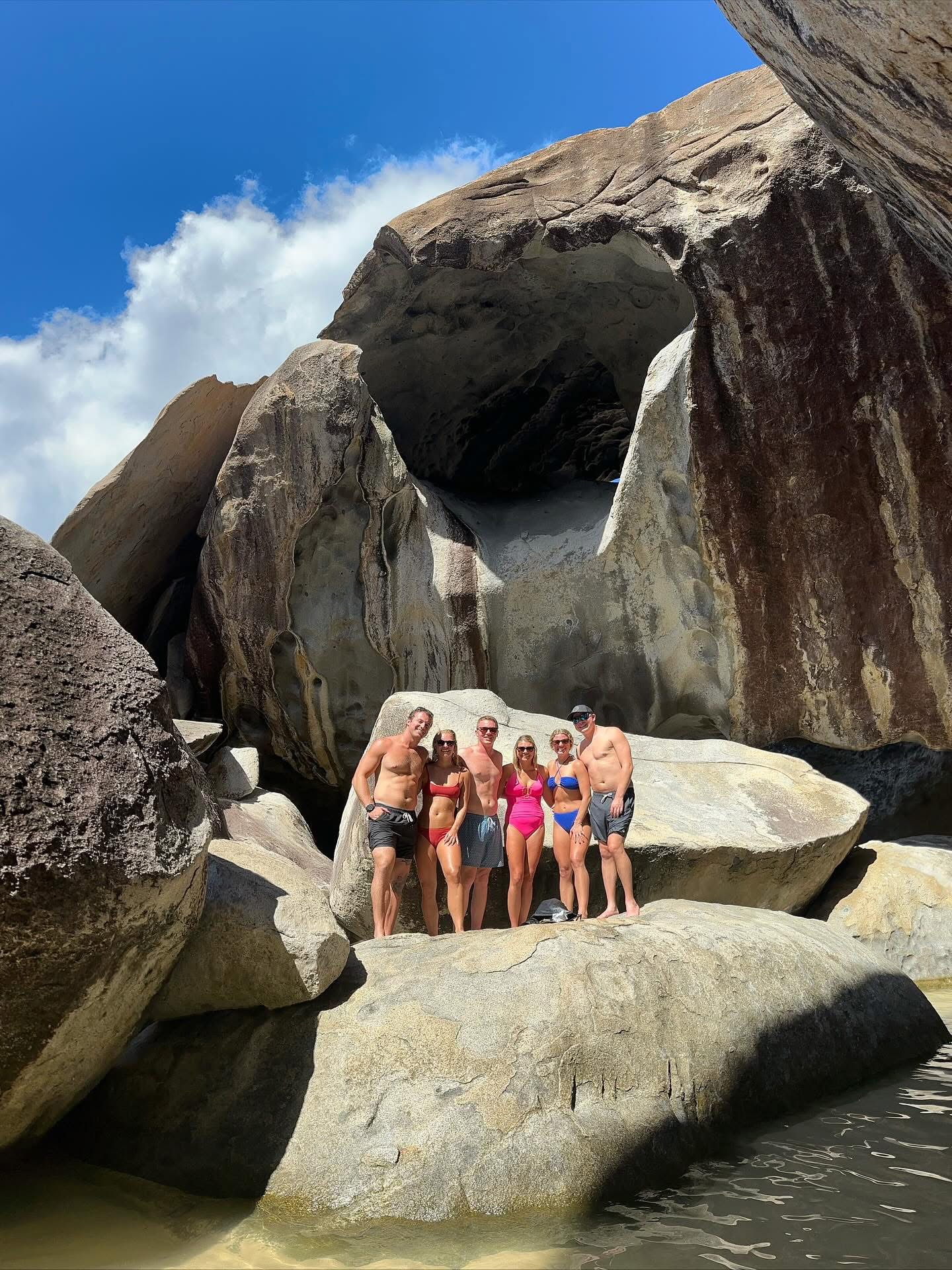 Six people in swimwear stand on rocks under a large cave-like formation.