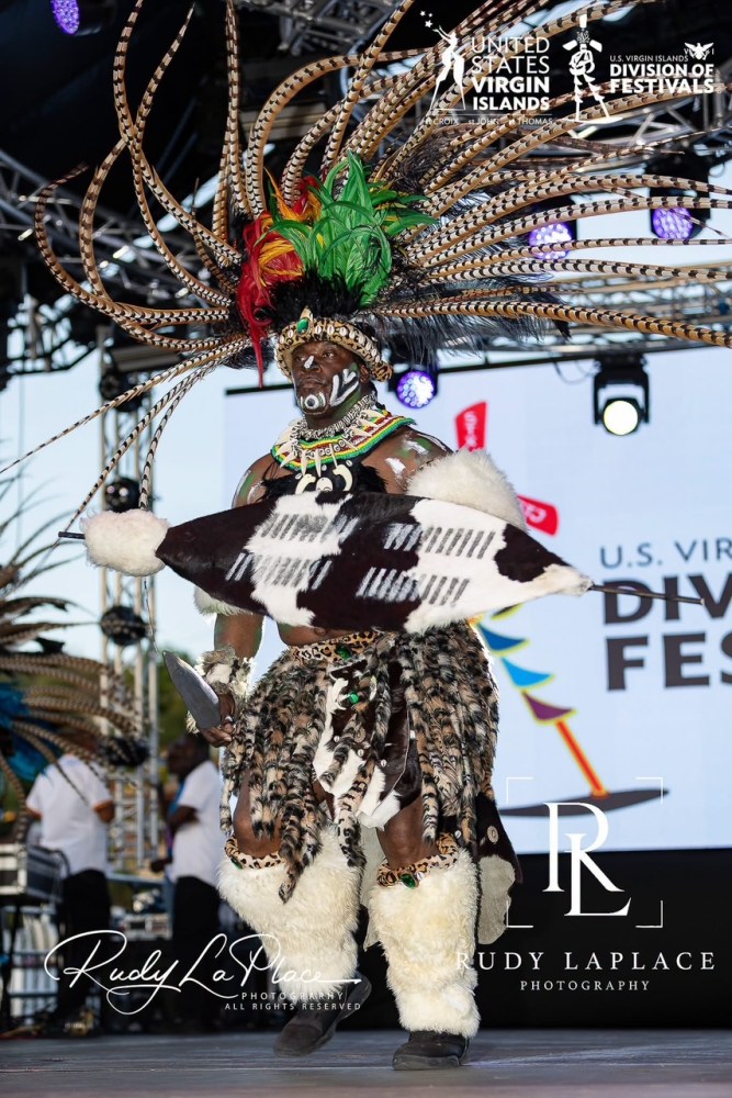 Person in elaborate costume with feathers and patterns performs on stage.