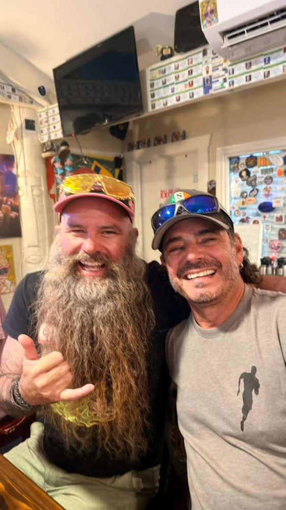 Two smiling men with beards, wearing caps and sunglasses inside a room with wall decorations.