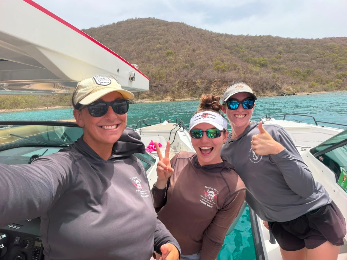 Three people in sunglasses smiling on a boat near a hilly shoreline.