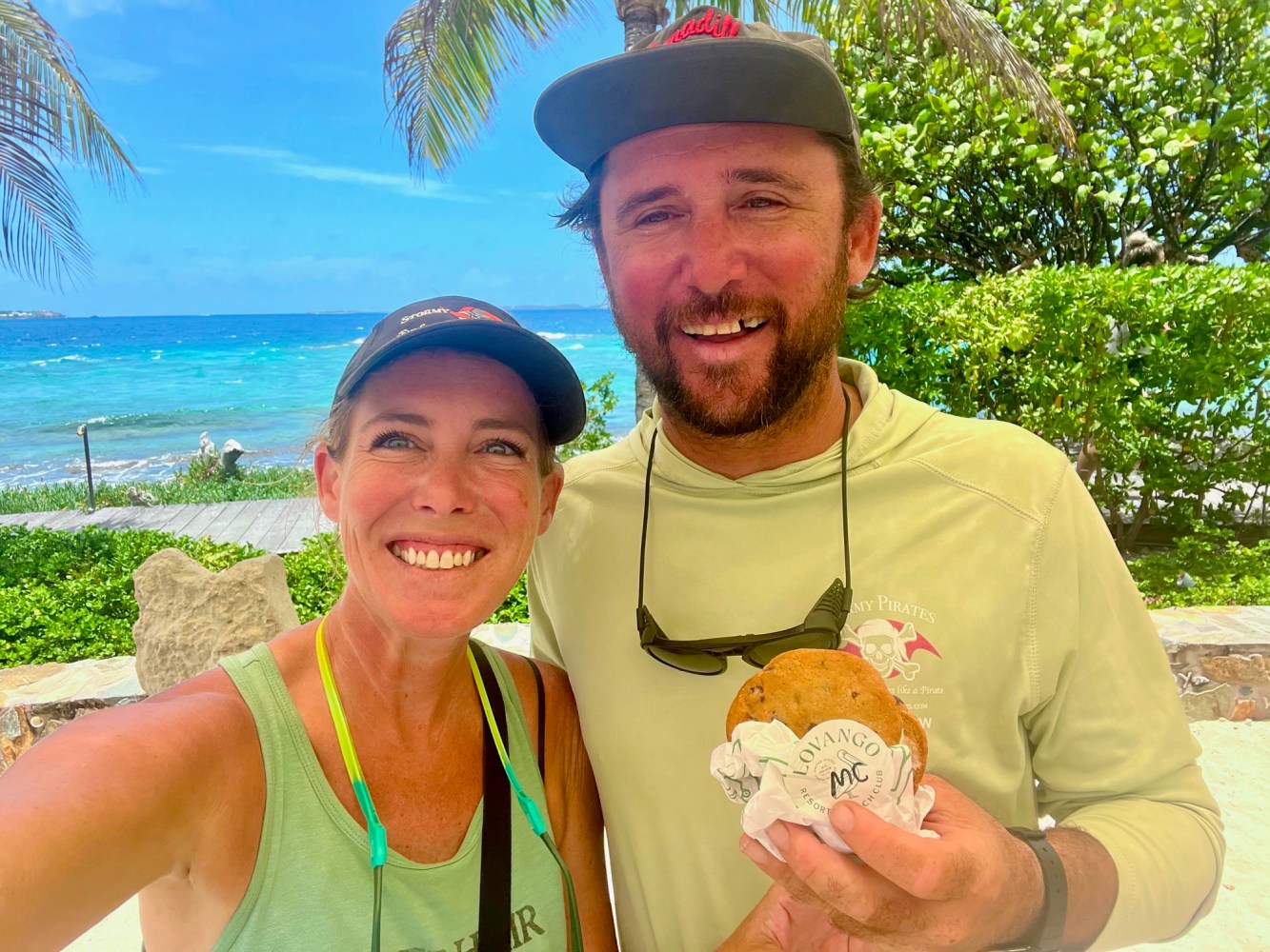 Two people smiling with cookies, palm trees and ocean in background.