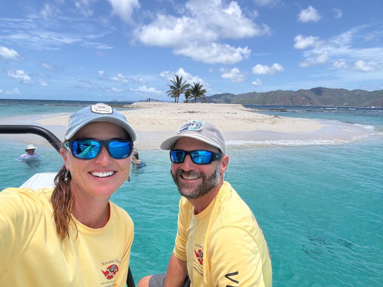 Two people in yellow shirts and caps smile on a boat near a sandy island with palm trees.