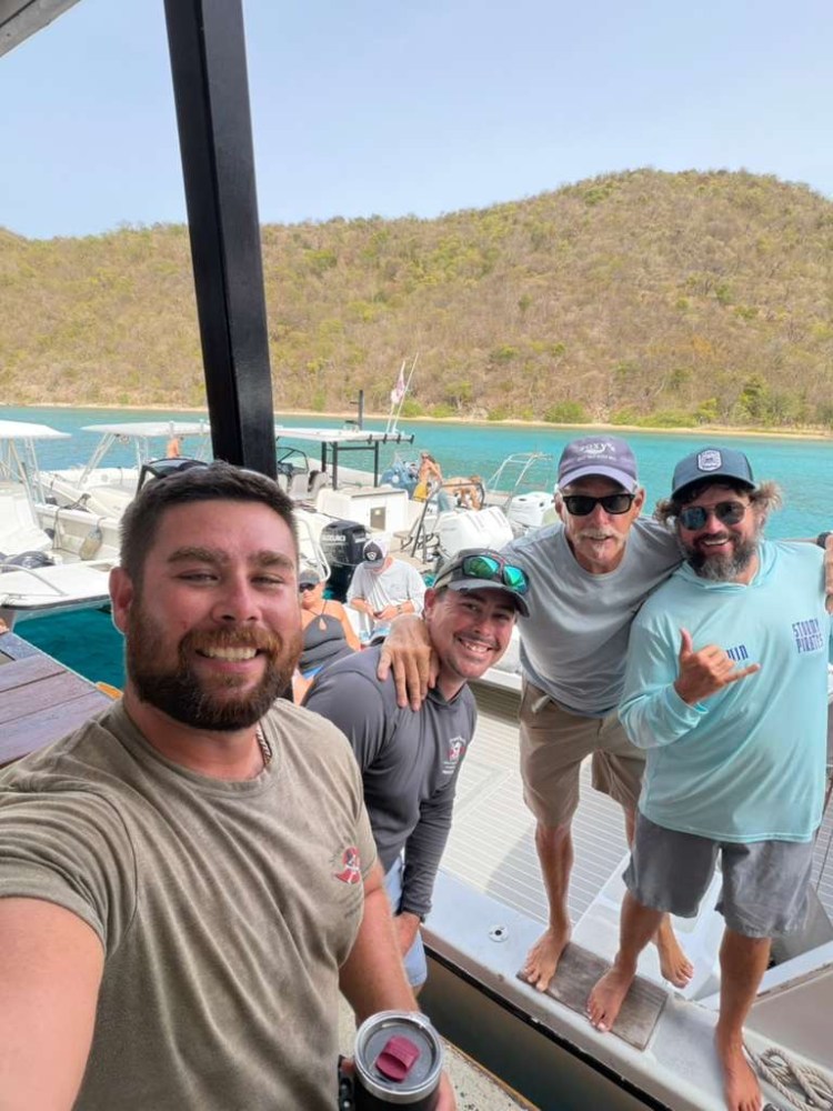 Four men smiling and posing on a dock with boats and a hilly landscape in the background.
